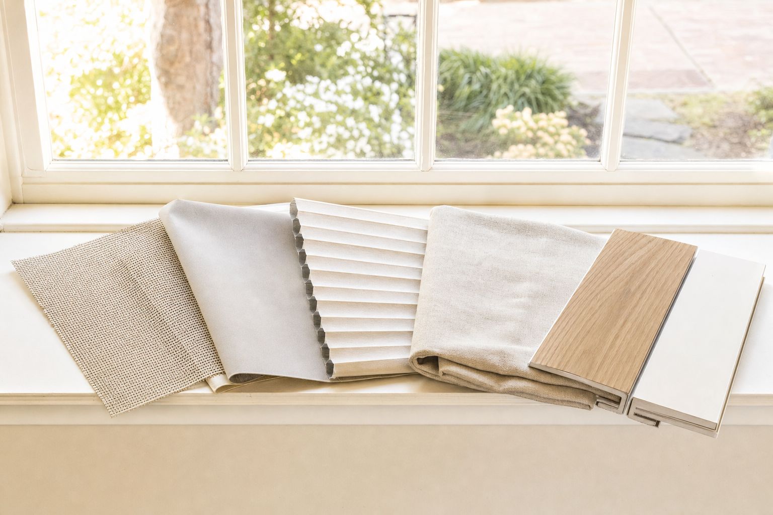 Window covering samples fanned along a sunlit windowsill — an open-weave solar screen glowing with daylight, a solid blackout swatch, a honeycomb cellular sample showing its cells, a folded length of oatmeal linen and two slats, one grained wood and one smooth white composite