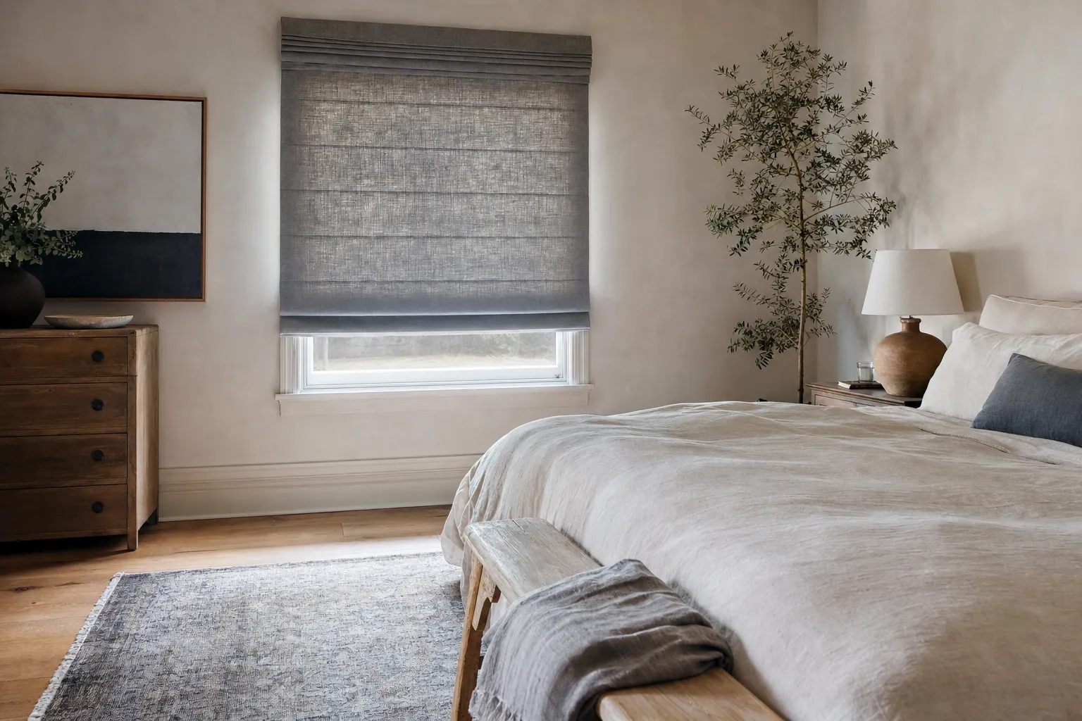 A lined roman shade lowered almost fully in a bright bedroom, the horizontal fold lines still readable across the cloth